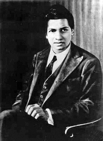 Photo of Srinivasa Ramanujan, sitting, wearing a dark suit and tie.